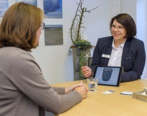 Beratungsgespräch zum Thema Waldbestattungen in einem Büro des Bremer Bestattungsunternehmens GE·BE·IN. Eine Mitarbeiterin zeigt einer Kundin eine Urne auf dem Bildschirm, im Hintergrund sieht man einen grünen Zweig.